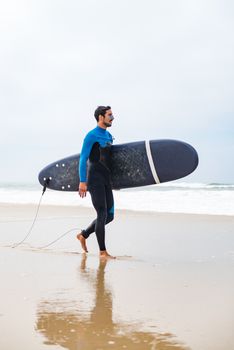 Young male surfer wearing wetsuit, holding surfboard under his arm, walking on beach after morning surfing session.