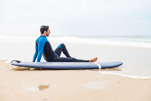 Young male surfer wearing wetsuit, sitting beside his surfboard on beach after morning surfing session.