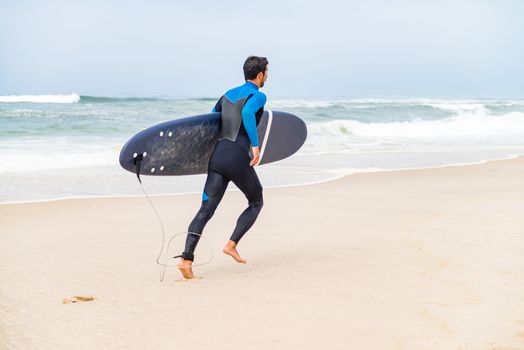 Young male surfer wearing wetsuit, holding surfboard under his arm, running on beach after morning surfing session.