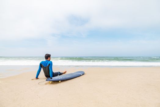 Young male surfer wearing wetsuit, sitting beside his surfboard on beach after morning surfing session.