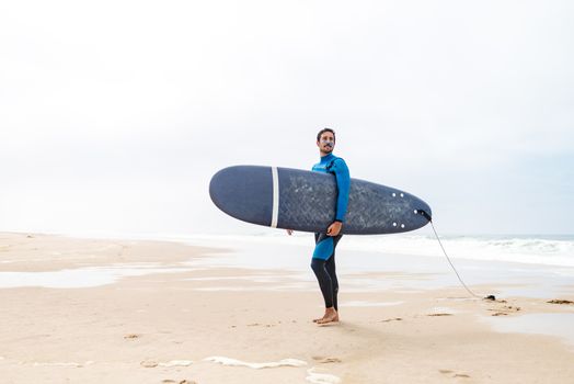 Young male surfer wearing wetsuit, holding surfboard under his arm, standing on beach after morning surfing session.