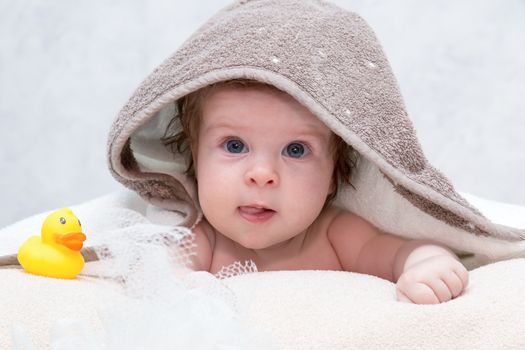 Baby girl under towel in bedroom after bath or shower. Yellow rubber duck and white washcloth lying near her. Textile and bedding for children