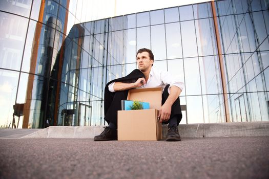 Fired business man sitting frustrated and upset on the street near office building with box of his belongings. He lost work