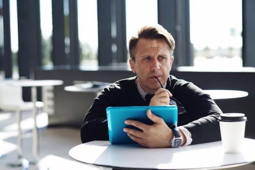 Portrait of thoughtful worried man sitting in cafe with digital tablet and biting glasses