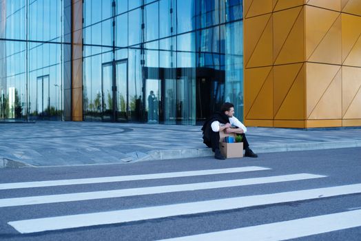 Fired business man sitting frustrated and upset on the street near office building with box of his belongings. He lost work