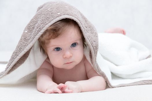 Cute newborn baby girl with beautiful blue eyes on a white terry coverlet. Adorable baby looking out under a white blanket or towel. Sad baby girl in white towel after shower