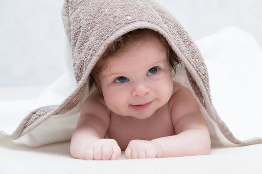 Smiling baby girl after shower or bath with towel on head. Happy laughing baby wearing hooded towel lying on parents bed after bath or shower. Clean dry child in bedroom. Children hygiene. Textile for infants. Looking up.
