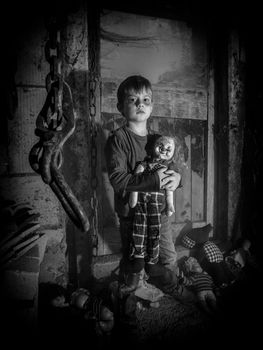 Photo of a creepy young boy holding an old clown doll in an old barn covered in spiderwebs and dust.