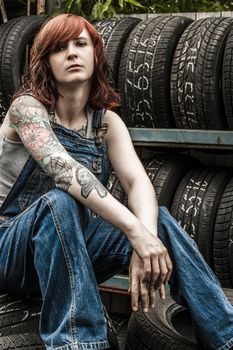 Photo of a young beautiful redhead mechanic wearing overalls and sitting behind an old garage. Attached property release is for arm tattoos.