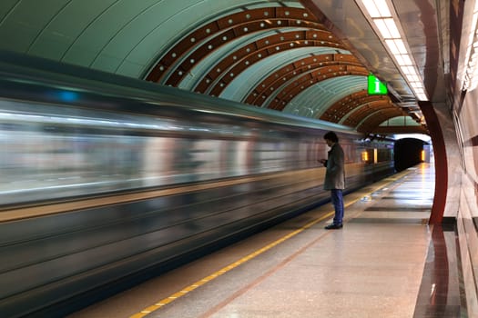 Lonely young man with smartphone shot from profile at subway station with blurry moving train in background