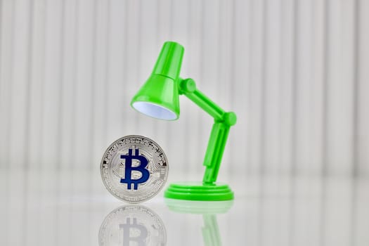 Digital currency physical silver bitcoin coin near green lamp on the table.