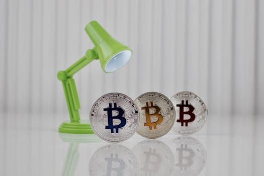 Cryptocurrency physical silver blue yellow and red bitcoin coin on white table near green lamp.