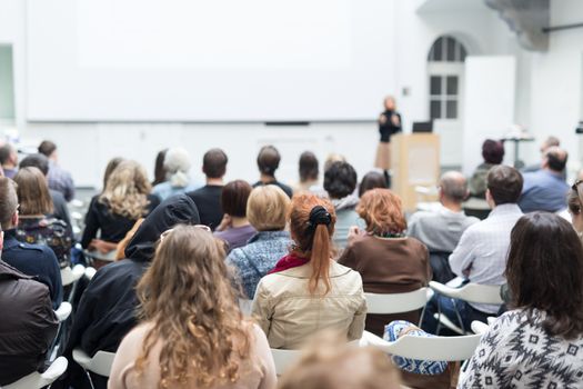 Business and entrepreneurship symposium. Female speaker giving a talk at business meeting. Audience in conference hall. Rear view of unrecognized participant in audience. Copy space on whitescreen.