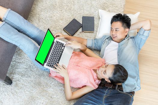 Young Asian Couples lying down and using laptop together in living room of contemporary house for modern lifestyle concept