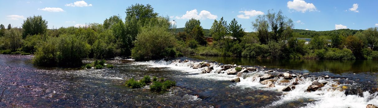 panoramic photo of a waterfall on a small river, with green trees on the opposite side