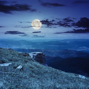 view on high mountains from hillside covered with grass with few stones on the edge at night in full moon light