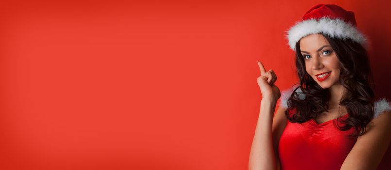 Young brunette christmas girl pointing at something on red background, copy space for text