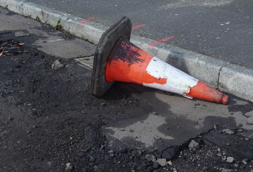 Orange and white damaged traffic cone lying in the gutter. The road surface is undergoing repairs with broken tarmac.