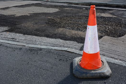 Bright orange and white traffic cone on a pavement, damaged road surface beyond, undergoing repair to the tarmac