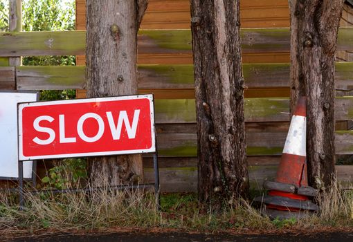 SLOW traffic sign with broken orange and white traffic cones, stored on the verge against tree trunks