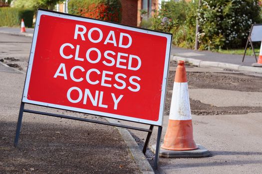 ROAD CLOSED ACCESS ONLY road sign on a pavement, with a traffic cone in the road. Broken tarmac surface visible in street beyond.
