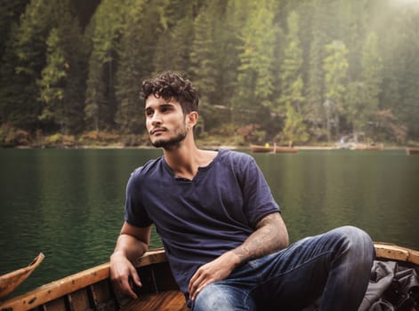 Handsome tattooed man sitting in boat on background of woods on shore of lake looking away.