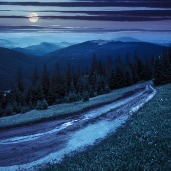 composite landscape with empty road to coniferous forest through the grassy hillside meadow on high mountain range at night in full moon light