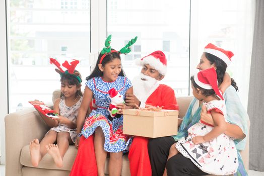 Happy Indian family celebrating Christmas holidays, with gift box sitting on sofa or couch at home, Asian people festival mood indoors. 