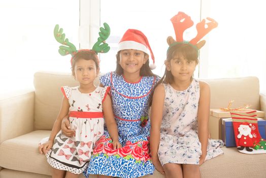 Happy Indian family celebrating Christmas holidays, with gift box and santa hat sitting on sofa or couch at home, adorable Asian children on festival mood. 