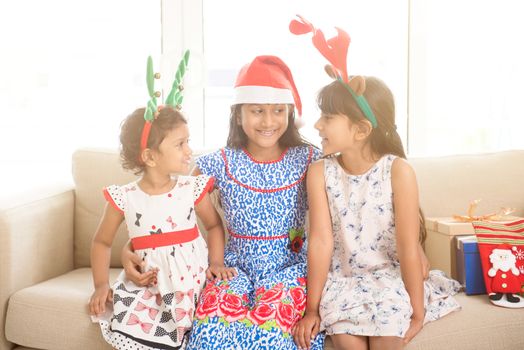 Happy Indian family celebrating Christmas holidays, with gift box and santa hat sitting on couch at home, adorable Asian children on festival mood. 