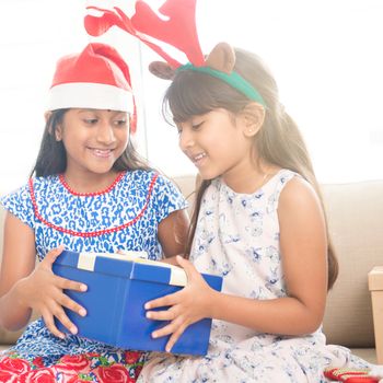 Happy Indian family celebrating Christmas holidays, with gift box and santa hat sitting on sofa at home, cute Asian children on festival mood. 