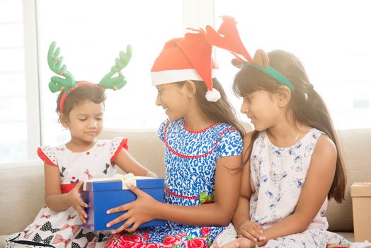 Happy Indian family celebrating Christmas holidays, with gift box and santa hat sitting on sofa at home, joyful Asian children on festival mood. 