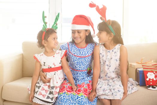 Happy Indian family celebrating Christmas holidays, with gift box and santa hat sitting on sofa at home, adorable Asian children on festival mood. 