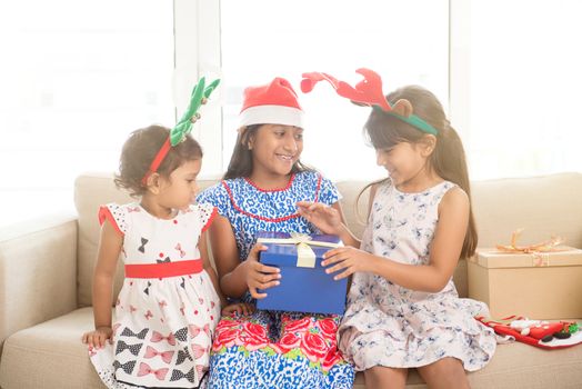 Happy Indian family celebrating Christmas holidays, with gift box and santa hat sitting on couch at home, cute Asian children on festival mood. 