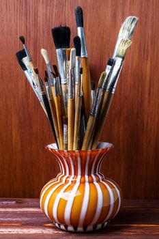 Assorted dirty brushes in a vase on a wooden background. Used artist paintbrushes in vase.