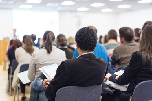 Business and entrepreneurship symposium. Female speaker giving a talk at business meeting. Audience in conference hall. Rear view of unrecognized participants making notes in audience.