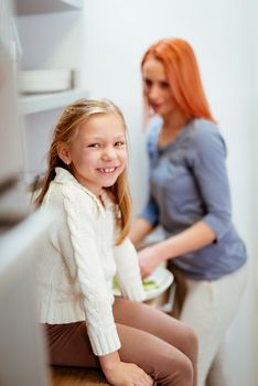 Cute smiling little girl and beautiful young woman preparing food in the kitchen. Looking at camera. Selective focus.