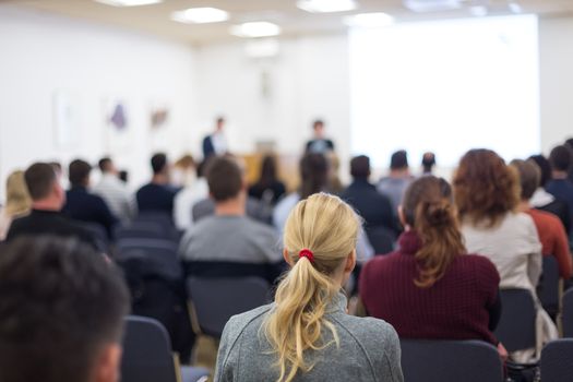 Business and entrepreneurship symposium. Female speaker giving a talk at business meeting. Audience in conference hall. Rear view of unrecognized participant in audience. Copy space on whitescreen.