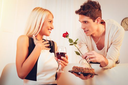 A young handsome man with a rose in his hand gives birthday cake to his beautiful surprised girl.