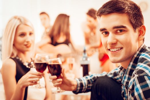 Handsome young man and his girlfriend toasting with red wine at the house party. Selective focus. Focus on guy. He is looking at camera.
