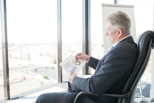 Mature businessman sitting alone in office with large panoramic windows with view at city and looking at diagrams and financial reports