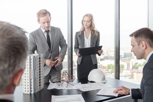 Business presentation of architects for investors, discussing new construction shapes, model of modern multi storey residential building house, blueprints and hardhat on table