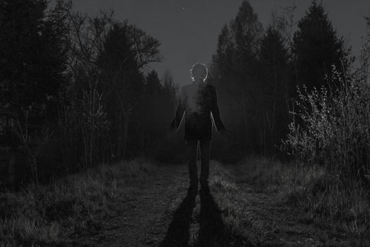 Male silhouette at the dark forest pathway through the bushes in the night. Man standing on the road against the car headlights. Mystery ghost concept. Black and white