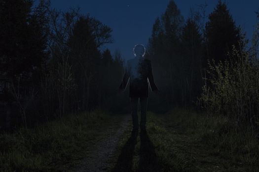 Male silhouette at the dark forest pathway through the bushes in the night. Man standing on the road against the car headlights. Mystery ghost concept. Soft focus.