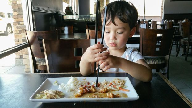 Cute Young Chinese and Caucasian Boy Learning To Use Chopsticks At Restaurant.