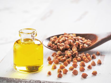 Brown buckwheat in spoon and buckwheat oil in glass bottle on white wooden background. Copy space.