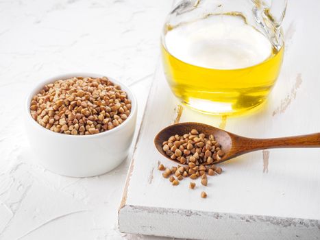 Brown buckwheat in spoon and buckwheat oil in glass bottle on white wooden background. Copy space.