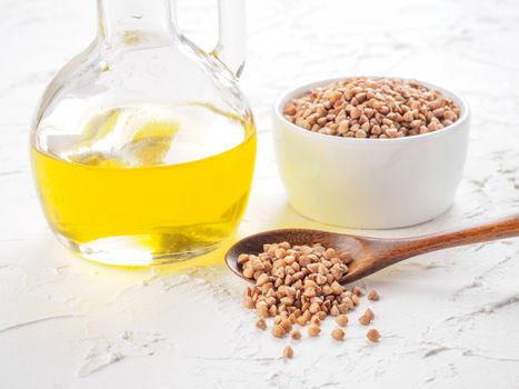 Brown buckwheat in spoon and buckwheat oil in glass bottle on white wooden background. Copy space.