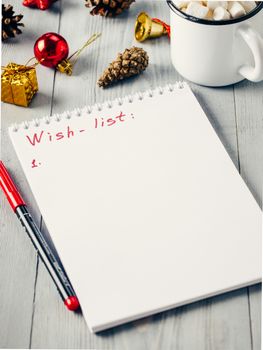 Christmas gifts shopping planning. Make shopping or to-do list for Christmas. Notebook, mug hot chocolate with marshmallows, New Year's decoration and pine cone on gray wooden background.