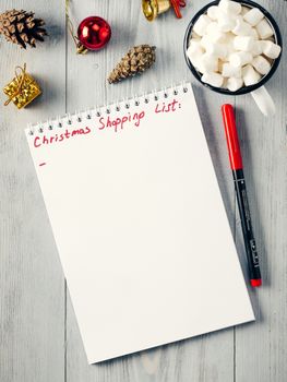 Christmas gifts shopping planning. Make shopping or to-do list for Christmas. Notebook, mug hot chocolate with marshmallows, New Year's decoration and pine cone on gray wooden background. Top view